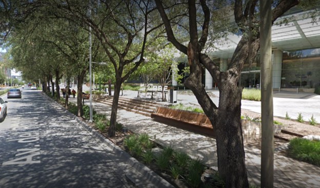 The image shows an avenue with cars driving and a sidewalk accessible to all users. The sidewalk has well-proportioned spaces. Comfortable wooden benches and stairs leading to a building can be seen. There is a lot of vegetation, with large, leafy trees providing shade to both the avenue and the sidewalk.