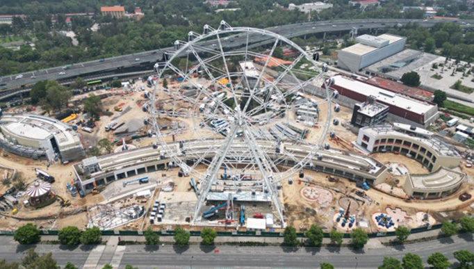 The image is shown in panoramic format, showing a land where an amusement park is being built. A large Ferris wheel of imposing dimensions is particularly prominent.