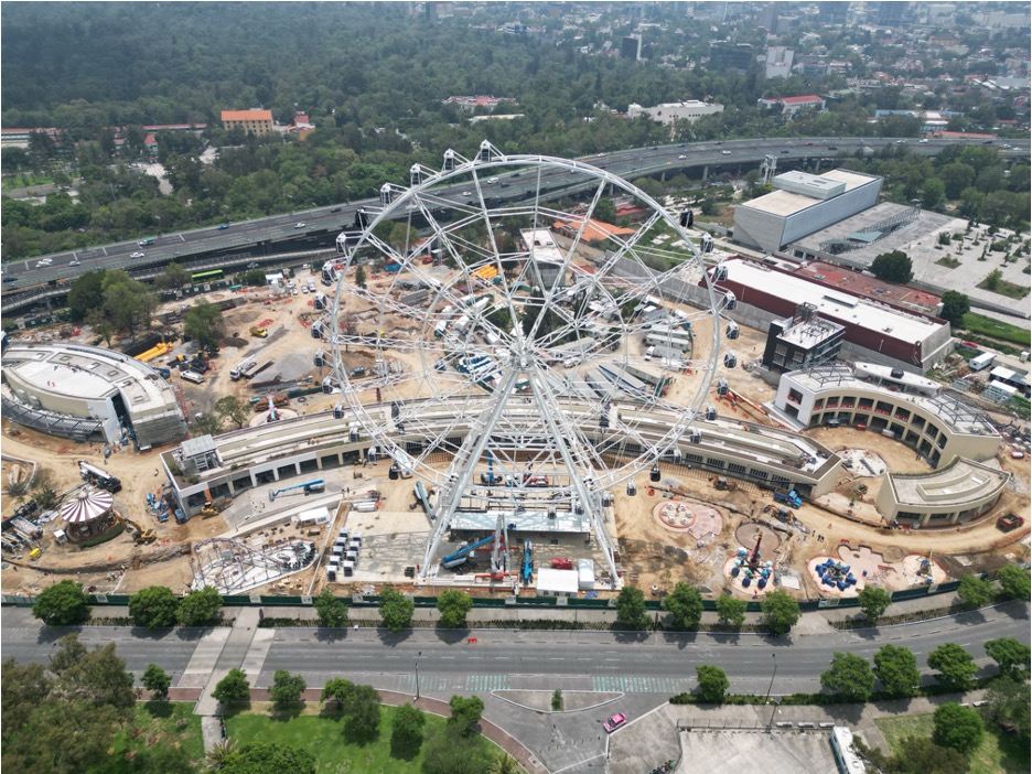 The image is shown in panoramic format, showing a land where an amusement park is being built. A large Ferris wheel of imposing dimensions is particularly prominent. The image shows a panoramic view of a large area with buildings. Four very large buildings surrounded by vegetation, trees, and pools stand out, giving it the appearance of a residential area. More buildings can be seen on the sides.