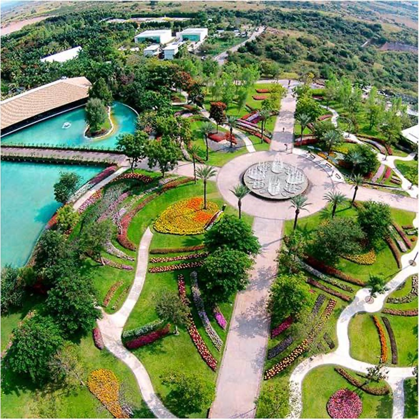 The image is shown in panoramic format and reveals a large area of vegetation. In the center of a large garden, a round fountain can be seen in operation. From the fountain, four paths lead to different gardens. On the right side, you can see a lake and a large bridge crossing it. The image shows a panoramic view of a large area with buildings. Four very large buildings surrounded by vegetation, trees, and pools stand out, giving it the appearance of a residential area. More buildings can be seen on the sides.