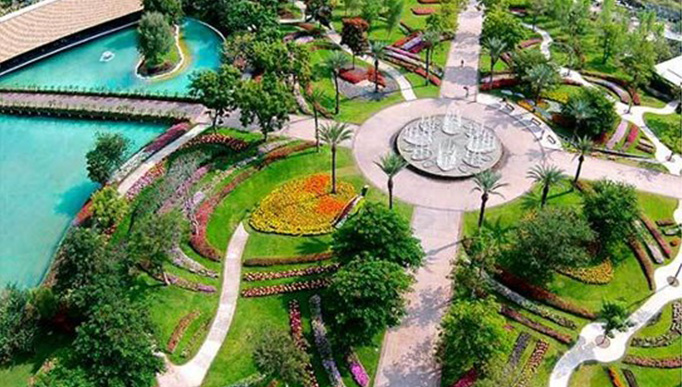 La imagen se muestra en formato panorámico y revela un extenso espacio de vegetación. En el centro de un amplio jardín se observa una fuente redonda en funcionamiento. Desde la fuente, se extienden cuatro pasillos que llevan a diferentes jardines. En la parte derecha, se puede ver un lago y un gran puente que lo cruza